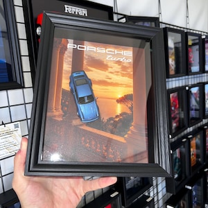 Peut inclure: Un cadre mural noir contenant une image d'une voiture miniature Porsche Turbo bleu clair garée sur un balcon surplombant une mer au coucher du soleil. Le texte PORSCHE turbo est imprimé en blanc en haut, devant des colonnes en pierre.