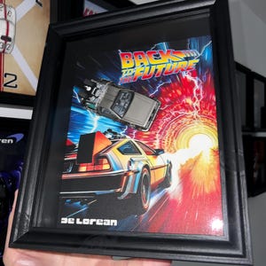 May include: Framed "Back to the Future" movie poster featuring a DeLorean time machine. The poster has a vibrant design with a red and orange tunnel effect, the DeLorean car, and the movie title in bold yellow and blue letters. The frame is black.