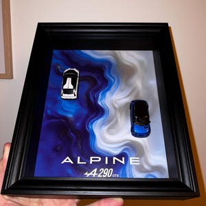Peut inclure: Présentation encadrée avec deux modèles réduits Alpine 4290 GTS. L'une est blanche avec des détails noirs, l'autre bleue. Le fond présente un motif abstrait bleu et blanc. Le cadre est noir et le texte "ALPINE 4290 GTS" est visible.