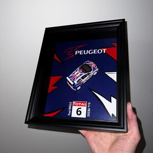 May include: A framed display featuring a miniature blue and white race car with the number 6 on the side. The car is on a blue background with a red and white design. The text "PEUGEOT" is at the top of the frame. The text "TOTAL" and "Paul Nagle" and "Kris Meeke" are on the bottom of the frame.
