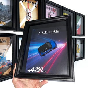 Peut inclure: Impression encadrée d'une voiture Alpine bleue, avec le texte "ALPINE" et "4290 GTS". L'impression est dans un cadre noir, avec d'autres impressions encadrées visibles. L'image est tenue par une personne.