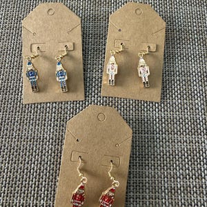 May include: Three pairs of nutcracker-shaped dangle earrings on brown cardboard display cards. One pair is blue and gold, one is white and gold, and one is red and gold. Each earring features a gold-tone hook and a metal nutcracker charm.