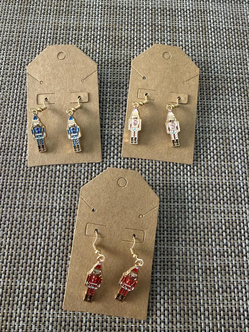 Puede incluir: Tres pares de pendientes colgantes con forma de cascanueces sobre tarjetas de cart&oacute;n marr&oacute;n. Un par es azul y dorado, otro blanco y dorado, y otro rojo y dorado. Cada pendiente tiene un gancho dorado y un dije de metal.