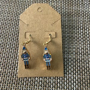 Puede incluir: Un par de pendientes colgantes de tono dorado con un dije de cascanueces. El cascanueces est&aacute; decorado con detalles de esmalte azul, blanco y dorado, incluyendo un sombrero azul, una chaqueta azul con detalles dorados y botas marrones.