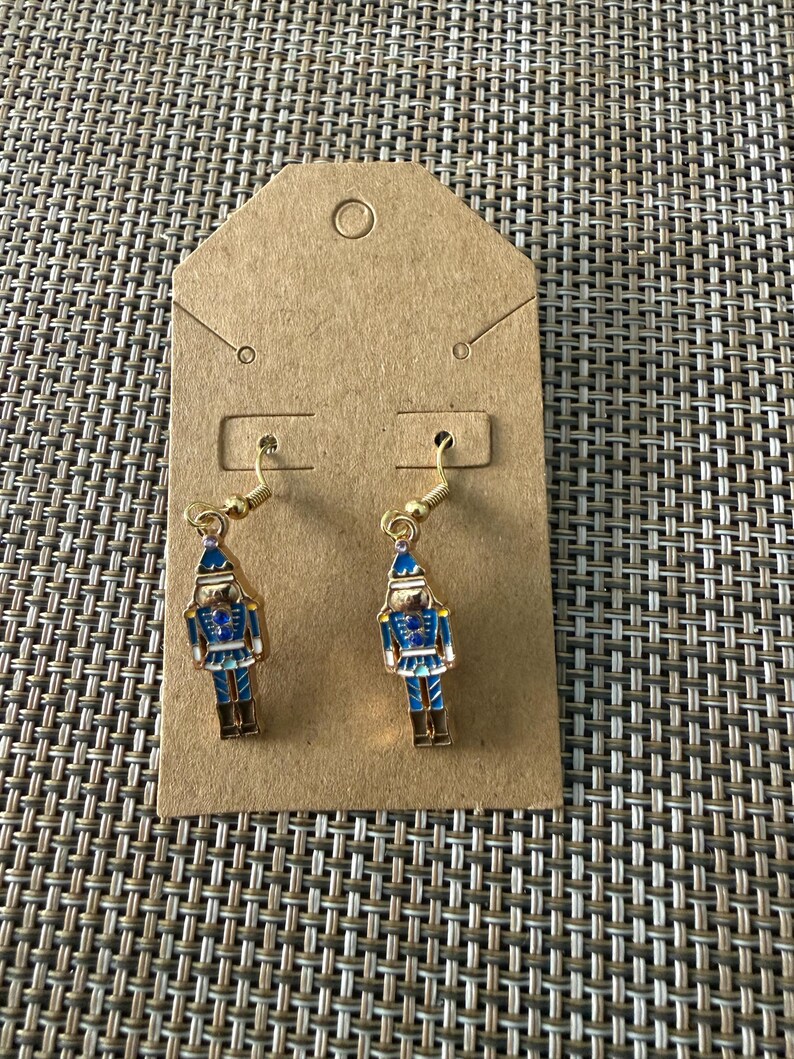 Puede incluir: Un par de pendientes colgantes de tono dorado con un dije de cascanueces. El cascanueces est&aacute; decorado con detalles de esmalte azul, blanco y dorado, incluyendo un sombrero azul, una chaqueta azul con detalles dorados y botas marrones.