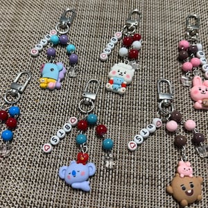 Op de afbeelding: Zes sleutelhangers met BT21-karakterbedels en letterkralen die BTS spellen met hartaccenten. Elke sleutelhanger bevat een zilverkleurige karabijnsluiting en kleurrijke ronde kralen. De karakters zijn onder andere Tata, Koya, RJ, Cooky, Shooky en Mang, elk met unieke kleurenschema's en kleine accessoires.