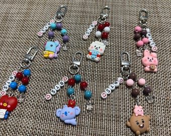 Puede incluir: Seis llaveros con dijes de personajes BT21 y cuentas de letras que deletrean BTS con detalles de corazones. Cada llavero incluye un cierre de mosquetón plateado y cuentas redondas de colores. Los personajes incluyen a Tata, Koya, RJ, Cooky, Shooky y Mang, cada uno con esquemas de color únicos y pequeños detalles.