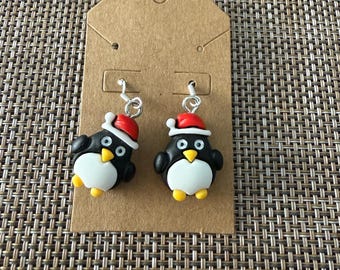 Puede incluir: Un par de pendientes colgantes con dijes de pingüino. Cada pingüino es blanco y negro con pico y patas amarillas, y lleva un gorro de Papá Noel rojo y blanco. Los dijes están sujetos a ganchos de metal de color plateado.