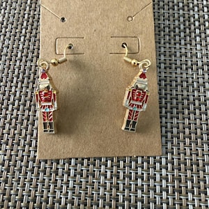 Puede incluir: Un par de pendientes colgantes de tono dorado con dijes de cascanueces. Cada dije est&aacute; detallado con esmalte rojo, blanco y negro y peque&ntilde;os diamantes de imitaci&oacute;n rojos en el pecho. Los pendientes cuelgan de ganchos de metal dorado.