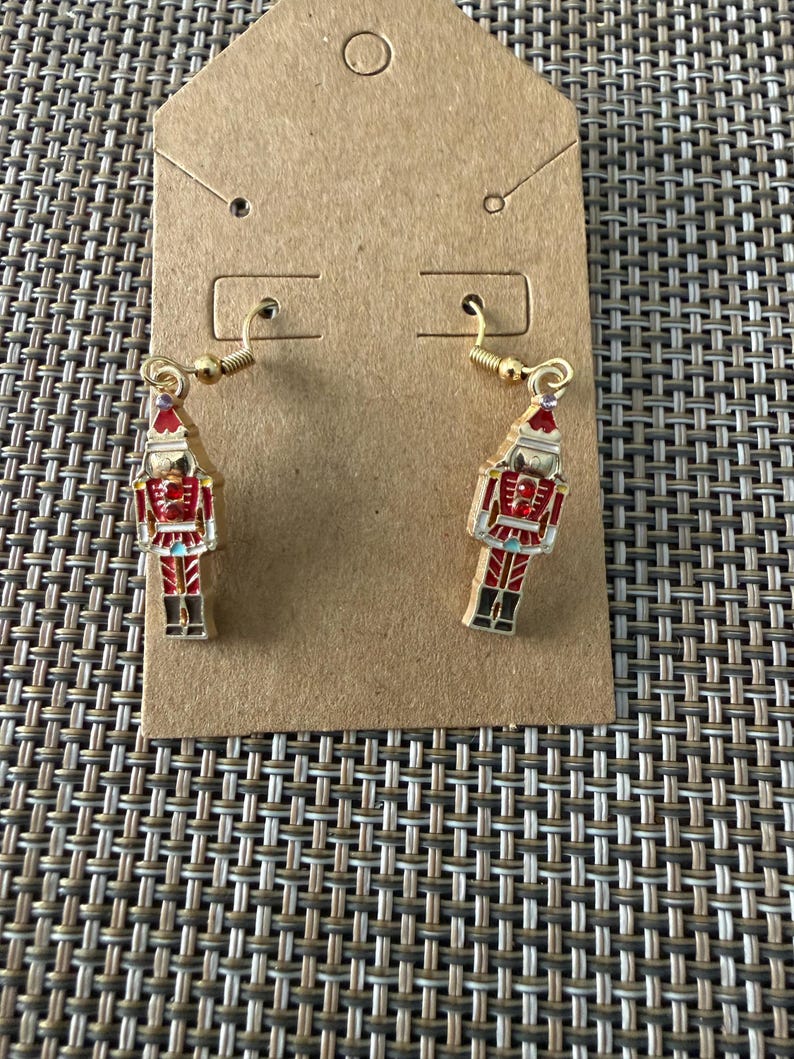 Puede incluir: Un par de pendientes colgantes de tono dorado con dijes de cascanueces. Cada dije est&aacute; detallado con esmalte rojo, blanco y negro y peque&ntilde;os diamantes de imitaci&oacute;n rojos en el pecho. Los pendientes cuelgan de ganchos de metal dorado.
