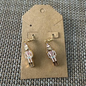Puede incluir: Un par de pendientes colgantes de tono dorado con un dise&ntilde;o de soldado cascanueces. Cada cascanueces est&aacute; acabado en esmalte blanco y rosa con peque&ntilde;os cristales rosas en el pecho y la cabeza, suspendidos de ganchos de metal dorado.