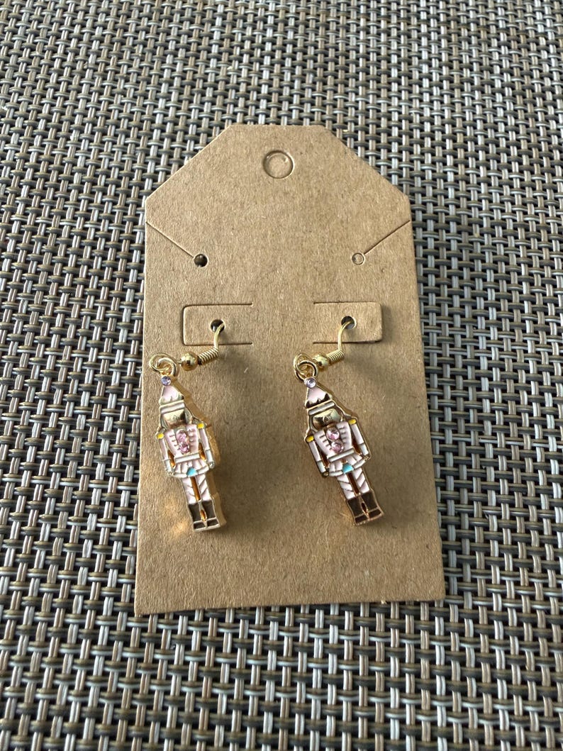 Puede incluir: Un par de pendientes colgantes de tono dorado con un dise&ntilde;o de soldado cascanueces. Cada cascanueces est&aacute; acabado en esmalte blanco y rosa con peque&ntilde;os cristales rosas en el pecho y la cabeza, suspendidos de ganchos de metal dorado.