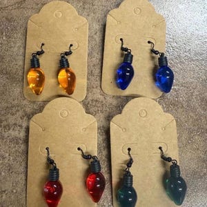 Op de afbeelding: Vier paar hangende oorbellen in de vorm van miniatuur kerstlampjes, elk met een doorschijnend oranje, blauw, rood of groen peertje, bevestigd aan zwarte metalen haakjes op individuele bruine kartonnen presentatiekaartjes.