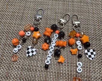Puede incluir: Tres llaveros con cierres de mosquetón plateados y cuentas variadas en naranja, negro y transparente. Los dijes incluyen un corazón a cuadros blanco y negro, un oso, una mariposa y cuentas de letras que forman LN, MCLAREN y OP 81.