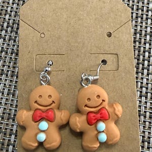 Op de afbeelding: Een paar bungelende oorbellen met lichtbruine peperkoekmannetjes. Elk figuurtje heeft een lachend gezicht, een rode strik en twee lichtblauwe ronde knoopjes op de borst. De bedels zijn bevestigd aan zilverkleurige metalen oorhaken.