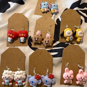 Op de afbeelding: Zeven paar bungelende oorbellen met kleurrijke cartoonfiguur-bedels op bruine kartonnen displaykaarten. De figuren omvatten rode harten, gele honden, bruine beren met roze konijntjes, witte schapen, lichtblauwe koala's en roze konijnen.
