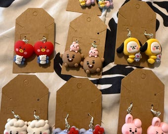 Puede incluir: Siete pares de pendientes colgantes con dijes de personajes de dibujos animados coloridos en tarjetas de presentación de cartón marrón. Los personajes incluyen corazones rojos, perros amarillos, osos marrones con conejitos rosas y koalas.
