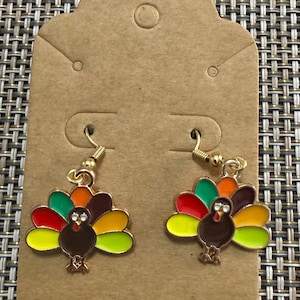 Op de afbeelding: Een paar goudkleurige bungelende oorbellen met emaille kalkoenbedels. Elke kalkoen heeft een bruin lichaam en een kleurrijke waaiervormige staart met rode, oranje, gele en groene segmenten. De oorbellen hangen aan goudkleurige oorhaakjes.