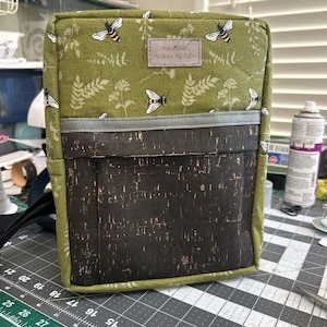 May include: A green fabric bag with a cork bottom and a zipper closure. The bag has a bee and floral pattern and the text "Handmade Creations By Salto" on the front.
