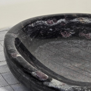 Könnte beinhalten: Eine dunkelgraue, oval geformte Schale mit polierter Oberfläche. Die Schale weist weiße und kastanienbraune Streifen im gesamten Stein auf. Die Schale steht auf einer grau-weiß karierten Oberfläche.