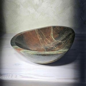May include: A handcrafted, oval-shaped stone bowl with a polished finish. The bowl features a blend of red, brown, and green hues with intricate veining patterns. It sits on a white wooden surface, showcasing its natural beauty and unique design.
