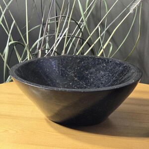 Op de afbeelding: Een zwarte, gespikkelde stenen kom met een breed, ondiep ontwerp. De kom staat op een lichte houten tafel, met lange, veervormige planten op de achtergrond. De binnenkant van de kom heeft een glad, gepolijst oppervlak.
