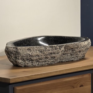 May include: A dark gray, oblong stone sink with a polished interior. The sink has a rough, textured exterior and is displayed on a wooden cabinet with a decorative drawer pull.