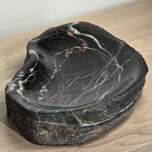 Op de afbeelding: Een zwarte marmeren zeepbakje met witte en koperkleurige aderen. Het bakje heeft een natuurlijke, organische vorm en staat op een houten oppervlak. Het is een decoratief item voor de badkamer of keuken.