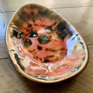 May include: A small, oval-shaped ceramic dish with a pink, black, and gold glaze. The dish has a slightly iridescent finish.