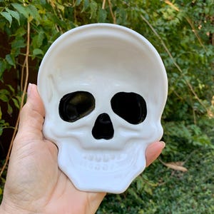 May include: A white ceramic skull-shaped dish featuring deep, glossy black indentations for the eye sockets and nasal cavity. The surface is smooth and reflective, with molded details defining the jawline and teeth structure.