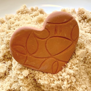 May include: A terracotta heart-shaped object with an etched design, resting on a bed of brown sugar. The heart has a smooth, matte finish and is a warm, earthy tone. The brown sugar provides a contrasting texture.
