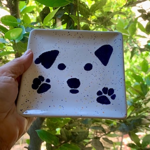 May include: Square white ceramic dish with a dog face design. The dog face is created with dark blue shapes and paw prints. The dish has a speckled pattern with small dots of color.