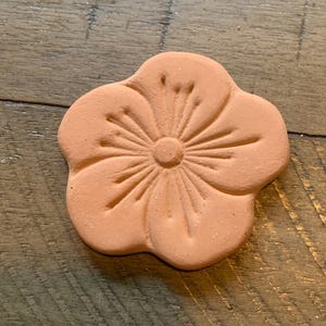 May include: A terracotta flower-shaped decorative piece. The flower has five rounded petals and a detailed center. The piece is a warm, earthy tone and sits on a wooden surface.