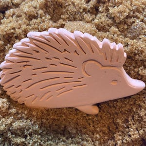 May include: A light pink ceramic hedgehog figurine with detailed etched spines. The hedgehog is lying down, with a small ear and a pointed snout. The figurine is set against a textured, brown background.