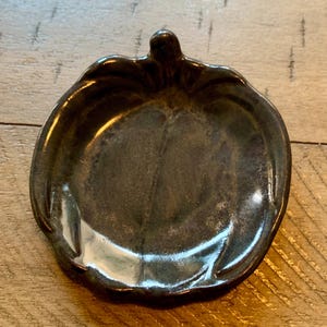 May include: A ceramic dish in the shape of a pumpkin, with a dark, glossy finish. The dish has a stem-like handle and a shallow bowl, perfect for serving small portions or as a decorative accent.