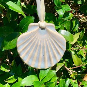 May include: A ceramic seashell ornament with a cream and light blue glaze, suspended by a cream-colored ribbon. The shell has a ridged design and is set against a backdrop of green leaves.