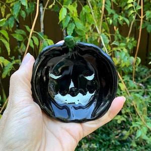 May include: A glossy black ceramic bowl shaped like a pumpkin, featuring a smooth, reflective surface and a small, distinct green stem at the top center. The bowl has a rounded, organic form with subtle indentations mimicking a pumpkin.