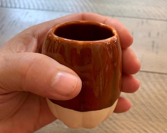 May include: A small, handmade ceramic cup with a glossy, brown glaze. The bottom portion of the cup is a light beige color. The cup is held in a person's hand.
