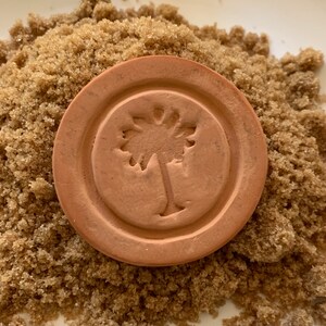 May include: A terracotta brown clay disc with a raised design of a palm tree. The disc is sitting on a pile of brown sugar.