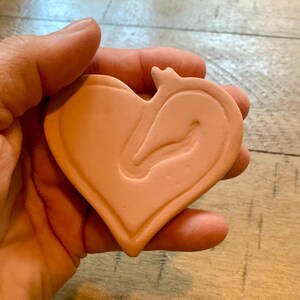 May include: A heart-shaped, terracotta-colored object with a raised, stylized design of a swan or bird. The object is held in a hand, showcasing its shape and texture. The design is centered within the heart outline.