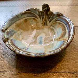 May include: A ceramic bowl with a pumpkin shape, featuring a blend of brown, tan, and light blue colors. The bowl has a glossy finish and a stem-like handle. The design is reminiscent of autumn and fall decor.