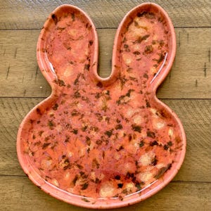 May include: A pink ceramic bunny-shaped dish with a speckled glaze. The dish has two ear-shaped indentations and a smooth, rounded bottom.