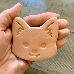 May include: A terracotta-colored ceramic fox head with a simple, hand-drawn design. The fox has closed eyes, a small nose, and a slight smile.