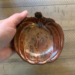 May include: A ceramic pumpkin-shaped dish with a textured, metallic finish. The dish is primarily brown with gold highlights, featuring a stem at the top. The design is reminiscent of autumn and fall decor.