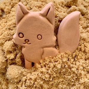 May include: A light brown fox-shaped object with a cartoonish design, featuring large round eyes and a fluffy tail. The fox is partially submerged in a bed of brown sugar, creating a playful and textured scene.