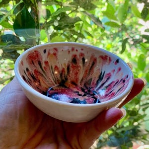 May include: A small, white ceramic bowl with a colorful interior design. The inside features a pattern of red, black, and orange hues. The bowl is held in a hand, with a blurred background of green foliage.