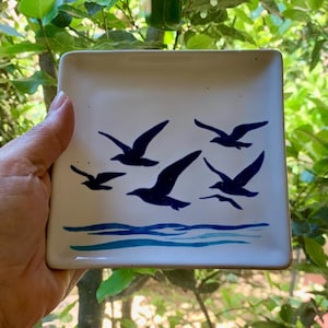 May include: Square white ceramic dish with a blue seagull design. The design features six dark blue seagulls flying above blue water lines. The dish is held in a hand, with a green leafy background.
