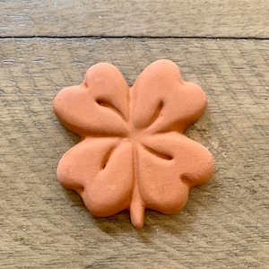 May include: A terracotta four-leaf clover charm. The charm is a light brown color and has a slightly textured surface.