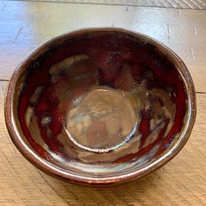 May include: A handmade ceramic bowl with a glossy, dark red interior and a bronze-colored rim. The bowl's surface reflects light, showcasing its unique glaze and texture. This piece is ideal for serving or as a decorative item.