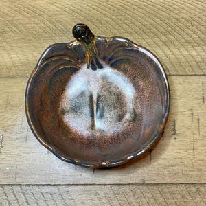 May include: A ceramic pumpkin-shaped dish with a stem. The dish has a brown and copper color scheme with a white center. The rim of the dish is slightly wavy, and the stem is dark brown.